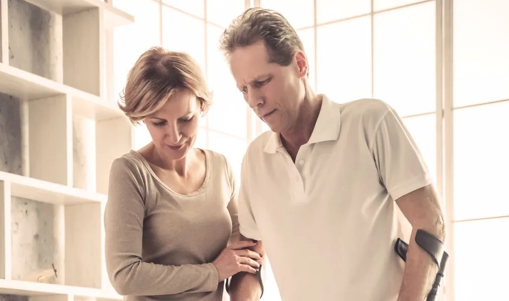 Zwei Personen stehen nah beieinander. Eine Person hält eine Krücke und scheint Unterstützung von der anderen Person zu erhalten, die einen Arm um den Rücken der ersten Person gelegt hat und deren Hand hält. Im Hintergrund ist ein großes Fenster mit natürlichem Licht und einige Regale auf der linken Seite des Bildes zu sehen.