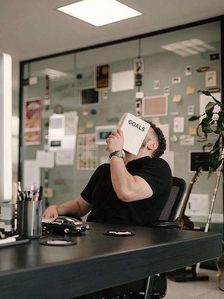 Ein junger Mann sitzt an einem Schreibtisch in einem modernen Büro und hält sich ein Buch vor das Gesicht. Seine Körperhaltung wirkt verzweifelt.