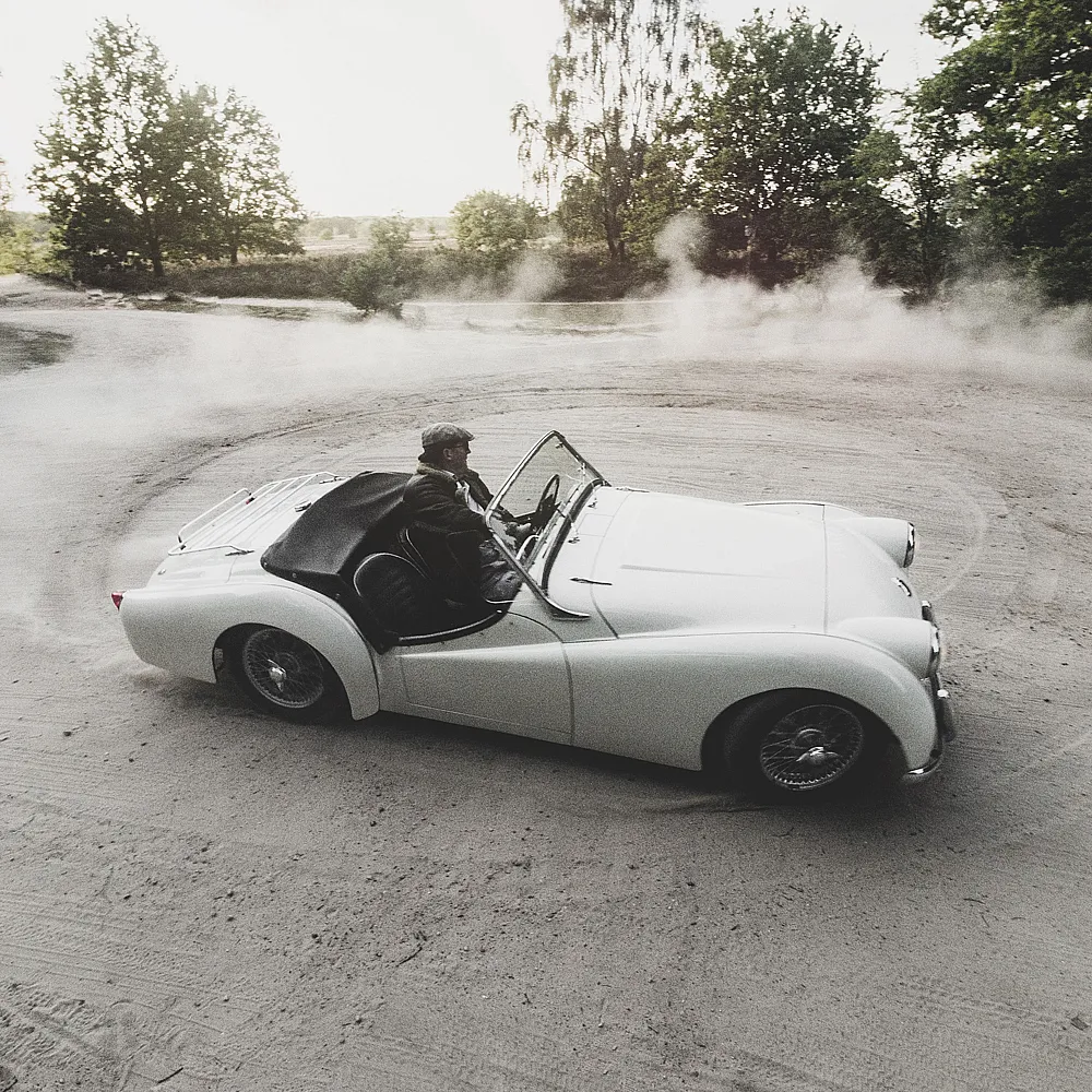 Ein weißes Cabrio-Oldtimer fährt auf einem Feldweg und wirbelt Staub auf. Der Fahrer trägt einen Hut. Im Hintergrund sind Bäume und Sonnenlicht zu sehen.