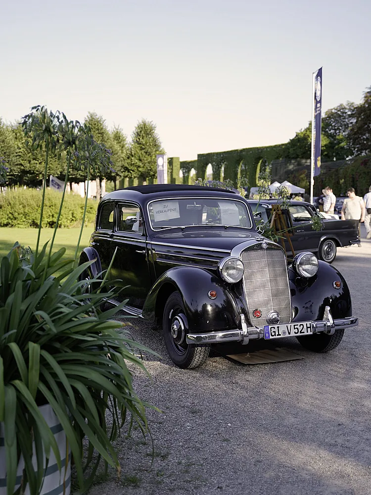 Ein schwarzer Mercedes-Benz Typ 170 V (W 136) steht vor einer Wiese, im Hintergrund sind weitere Fahrzeuge und Personen erkennbar