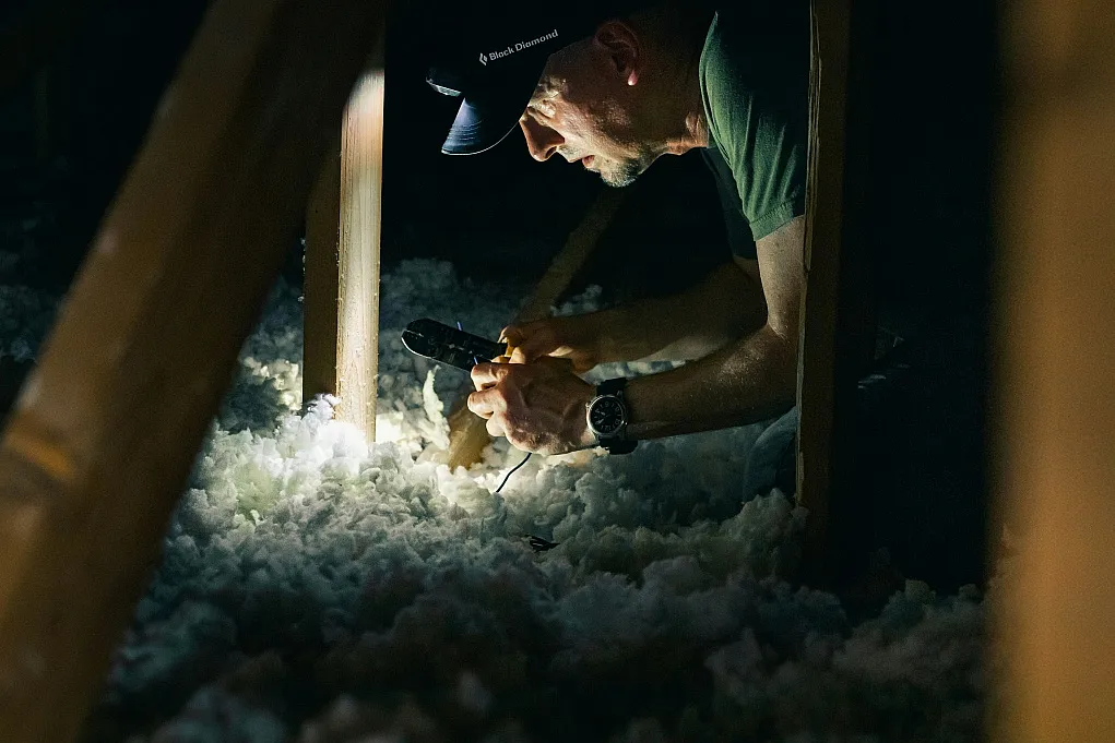 Fotografie zeigt Mann mit Taschenlampe in dunklem Dachboden, der Isoliermaterial inspiziert. Fokus liegt auf sorgfältiger Untersuchung und handwerklicher Arbeit in beengtem Raum mit Holzträgern.