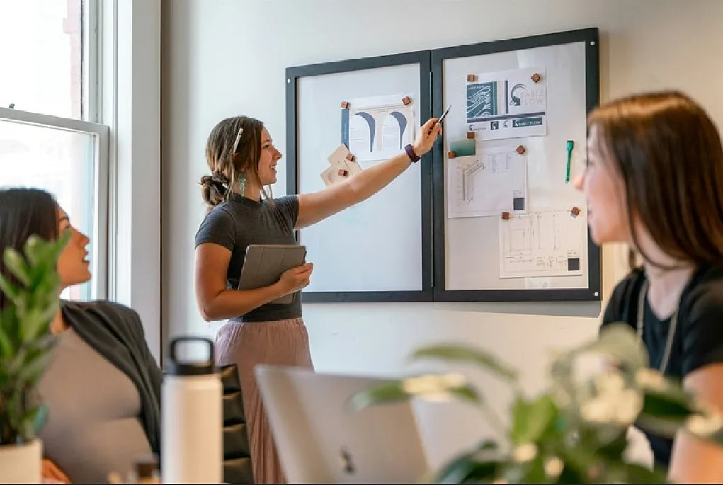 Fotografie einer Geschäftspräsentation in einem Büro mit drei Frauen, eine zeigt Diagramme und Grafiken an zwei Pinnwänden. Fokus liegt auf Zusammenarbeit und visueller Darstellung von Daten, um Strategien oder Ergebnisse zu erläutern.