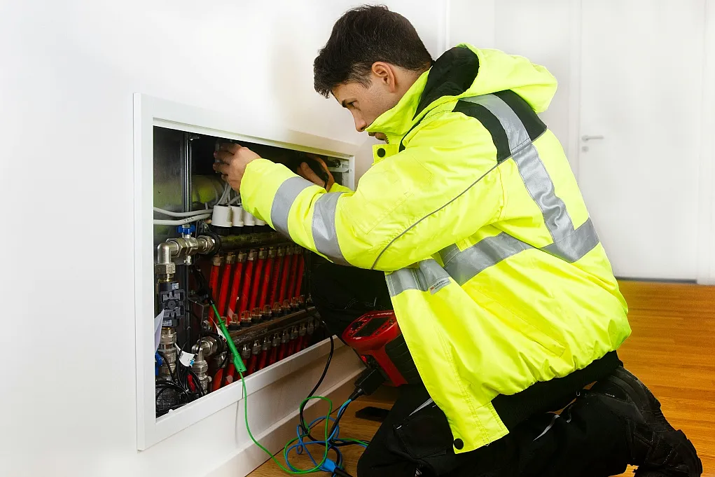 Fotografie eines Technikers in gelber Warnjacke, der an einem geöffneten Heizungsverteilerkasten in einem Innenraum arbeitet. Fokus liegt auf den roten und silbernen Ventilen sowie den angeschlossenen Kabeln, die auf Wartungs- oder Reparaturarbeiten hinweisen.