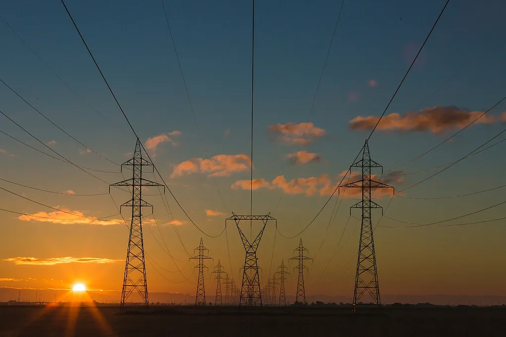 Fotografie von Hochspannungsmasten bei Sonnenuntergang mit orangefarbenem Himmel und vereinzelten Wolken. Stromleitungen verlaufen symmetrisch von vorderen zu hinteren Masten, was Tiefe und Perspektive betont.