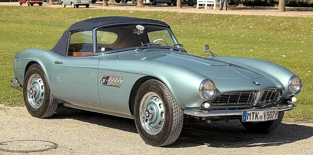Fotografie eines klassischen BMW 507 Roadsters in hellblau mit schwarzem Verdeck, geparkt auf einem gepflasterten Weg vor einer Grünfläche. Fahrzeugdetails wie runde Scheinwerfer, Chromstoßstangen und ein deutsches Kennzeichen sind gut sichtbar, was den eleganten Stil und den historischen Wert des Oldtimers betont.