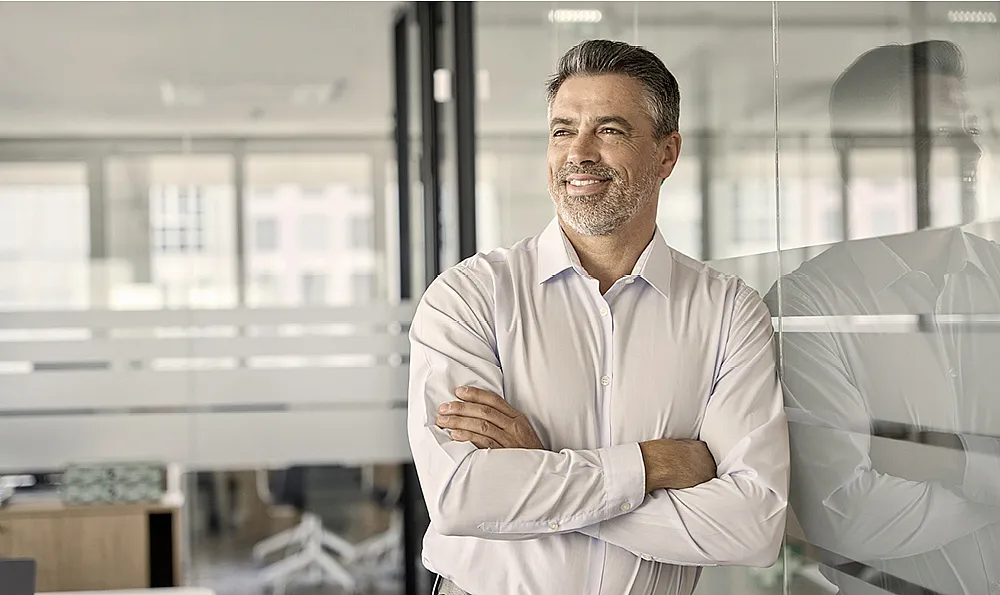 Ein Mann mit grauen Haaren und Bart, der ein helles Hemd trägt, steht lächelnd mit verschränkten Armen in einem modernen Büro.