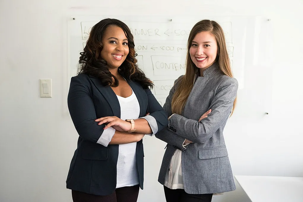Fotografie von zwei Frauen in Business-Kleidung, die selbstbewusst mit verschränkten Armen vor einem Whiteboard stehen. Szene vermittelt professionelle Zusammenarbeit und positive Arbeitsatmosphäre in einem modernen Büro.
