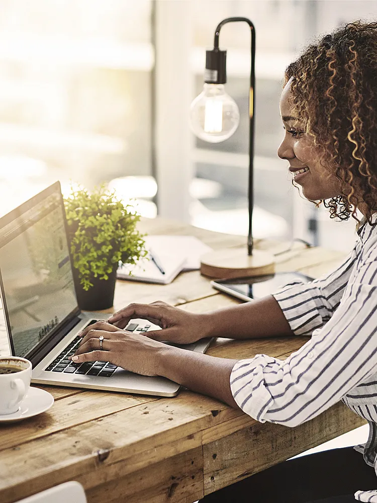 Eine Frau mit Locken und einer gestreiften Bluse sitzt am Schreibtisch und arbeitet an ihrem Laptop