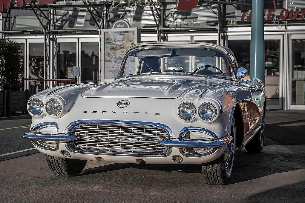 Fotografie eines klassischen Chevrolet Corvette Sportwagens in weiß mit Chromdetails, aufgenommen vor einem Restaurant. Fokus liegt auf Frontansicht mit markantem Kühlergrill, Doppel-Scheinwerfern und glänzender Lackierung bei Tageslicht.