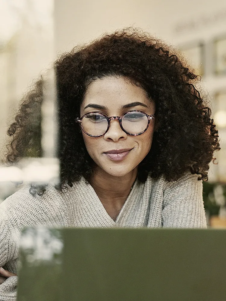 Eine Frau mit Locken und eine Brille blickt lächelnd auf den Bildschirm Ihres Laptops