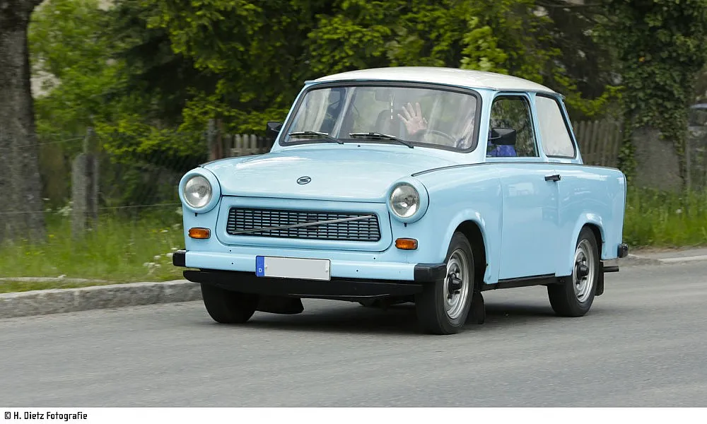 Ein hellblauer Oldtimer Trabant 601 mit weißem Dach fährt auf einer Straße, im Hintergrund ist grüne Vegetation zu sehen – das Fahrzeug bewegt sich nach links.