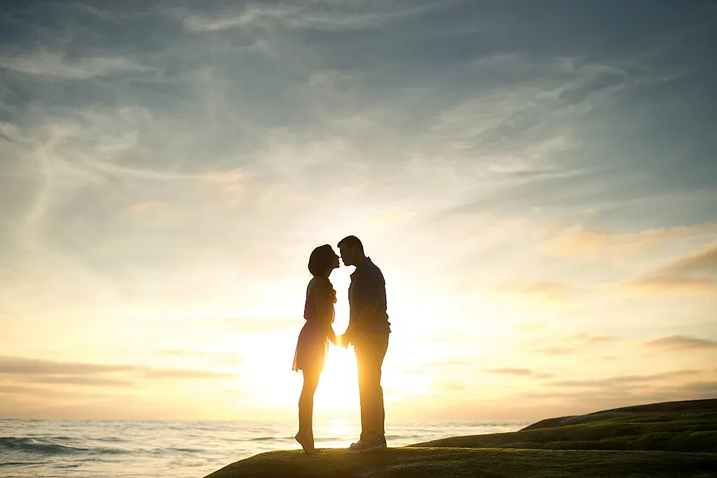 Fotografie zeigt Silhouetten eines Paares, das sich bei Sonnenuntergang am Meer nahekommt. Himmel mit sanften Wolken und warmem Licht erzeugt romantische Atmosphäre, während Felsen im Vordergrund ruhige Szene ergänzen.
