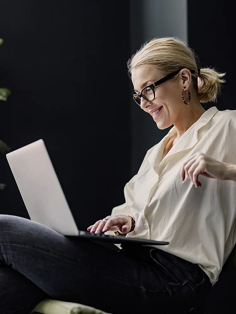 Eine Frau mit Brille und weißer Bluse sitzt auf einem Stuhl und blickt lächeln auf den Bildschirm ihres Laptops, der auf ihrem Schoß ist