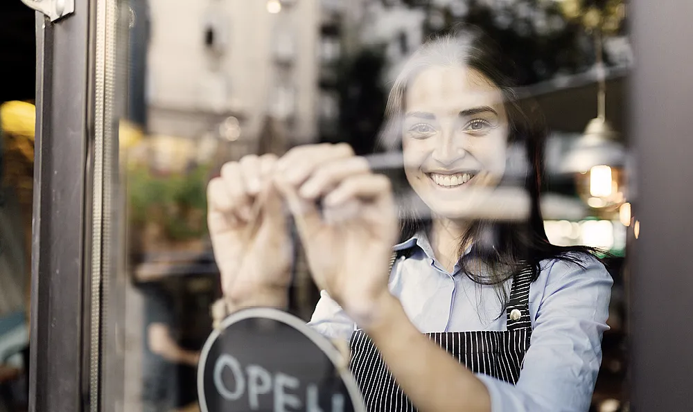 Eine lächelnde Frau in einer gestreiften Schürze öffnet ein Café und richtet ein Schild an der Glastür aus. Die Szene ist warm und einladend, mit verschwommener Straßenansicht draußen.