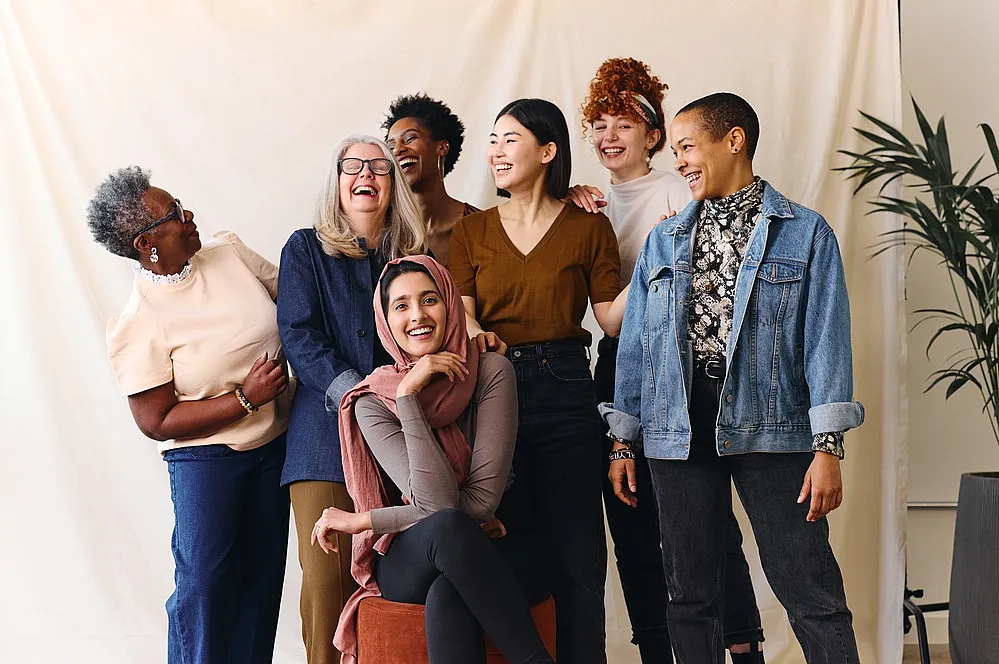 Sieben Frauen unterschiedlicher Herkunft stehen zusammen, lächeln und lachen. Sie strahlen in einer ungezwungenen, freundlichen Umgebung ein Gefühl der Zusammengehörigkeit und Freude aus. Sie repräsentieren gründende Frauen.