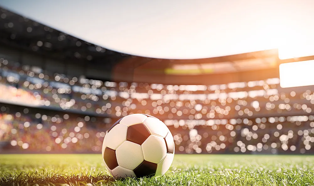 Ein Fußball liegt auf einem grünen Spielfeld in einem Stadion, das von Sonnenlicht beleuchtet wird. Im Hintergrund sind unscharfe Tribünen mit vielen Zuschauern zu sehen.