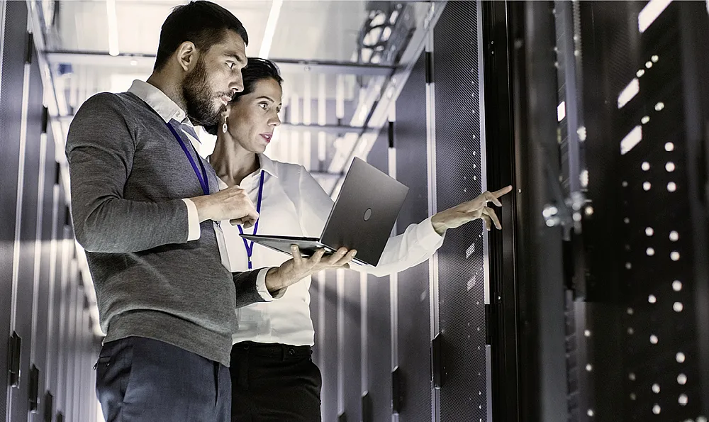 Zwei Personen in einem Serverraum, eine Person hält einen Laptop und die andere zeigt auf ein Server-Rack. Beide tragen ID-Karten um den Hals.
