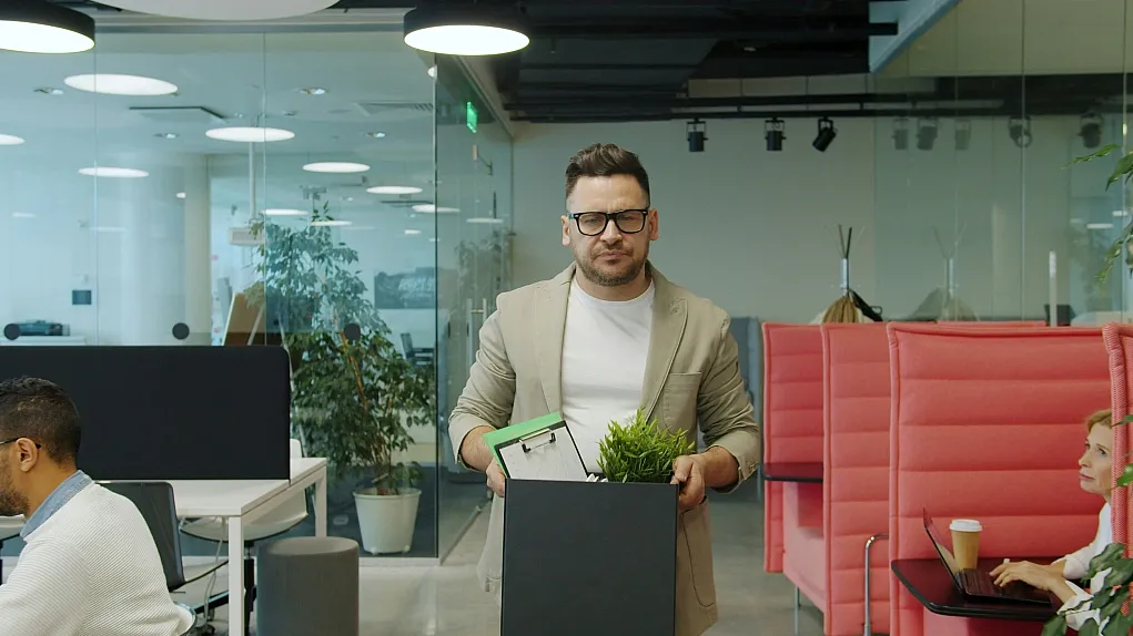 Fotografie eines Mannes, der ein schwarzes Karton mit persönlichen Gegenständen wie einer Pflanze und einem Ordner in einem modernen Büro trägt. Im Hintergrund sind mehrere Personen an Schreibtischen und rote Sitznischen mit Arbeitsplätzen sichtbar, was eine Büroumgebung mit offener Raumgestaltung zeigt.