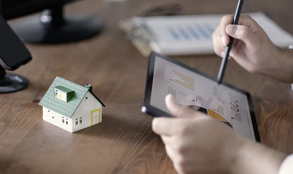 Eine Person arbeitet mit einem Stift auf einem Tablet, auf dem Diagramme angezeigt werden, daneben steht ein kleines Modellhaus auf einem Holztisch. Die Atmosphäre ist konzentriert und analytisch.