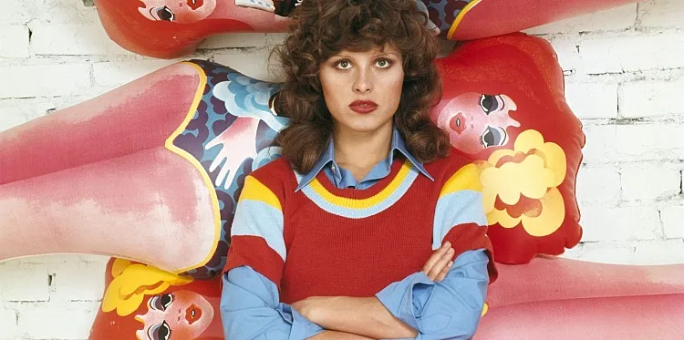 Eine Person mit lockigem Haar und ernstem Gesichtsausdruck steht vor einer Backsteinmauer, umgeben von bunten aufblasbaren Comicfiguren. Der Stil ist skurril und im Retro-Look gehalten.