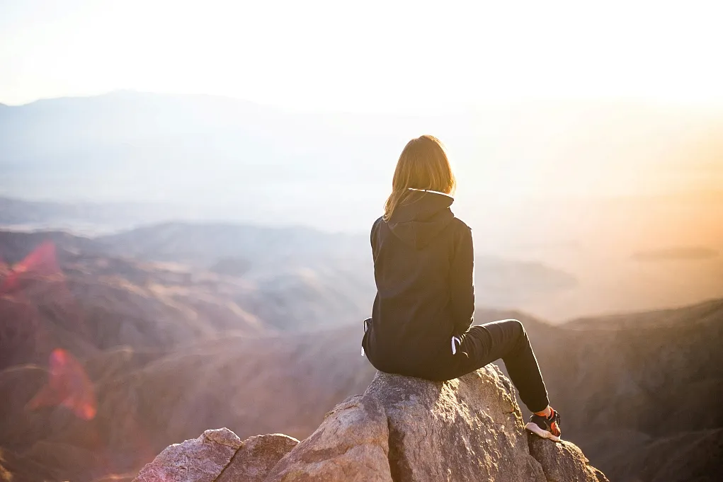 Fotografie einer Person mit Kapuzenjacke, die auf einem Felsen sitzt und in eine weite, bergige Landschaft bei Sonnenaufgang oder Sonnenuntergang blickt. Die Szene vermittelt Ruhe und Weite, mit warmem Licht und sanften Farben, die Atmosphäre von Naturerlebnis und Einsamkeit betonen.