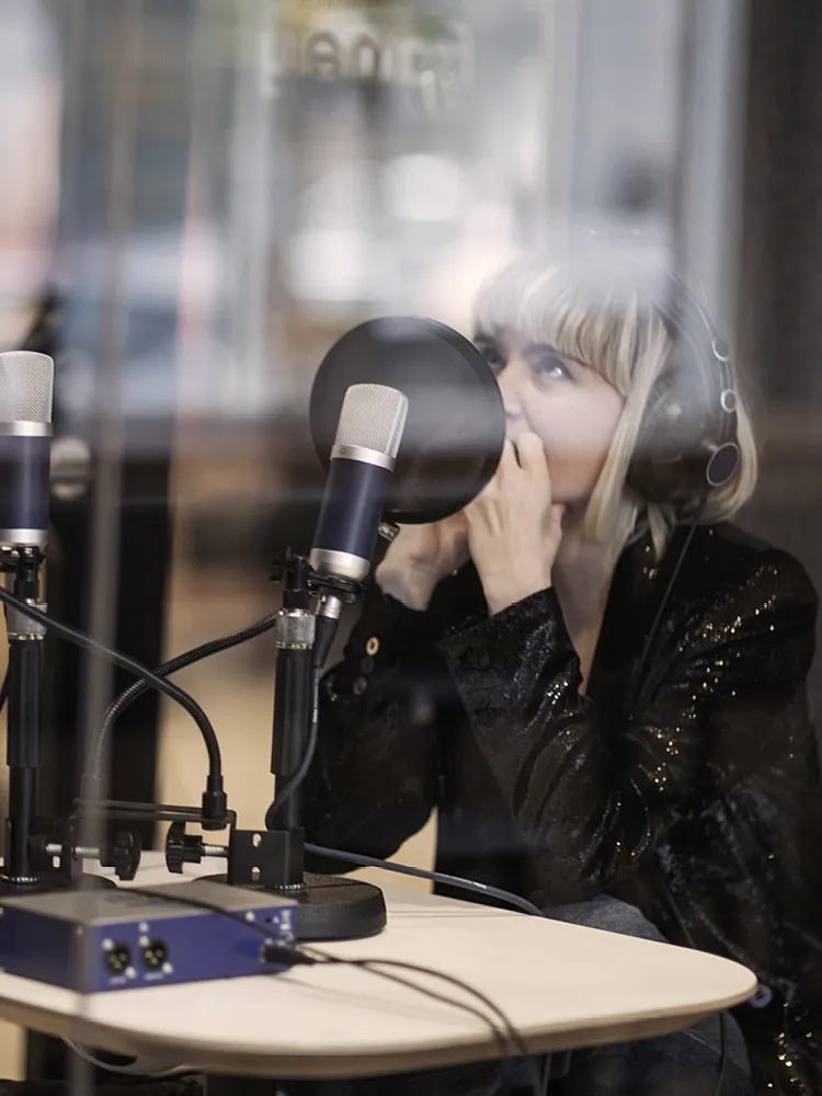 Frau mit blonden Haare und Lederjacke sitzt vor eine Podcast-Mikrofone und trägt Kopfhörer.