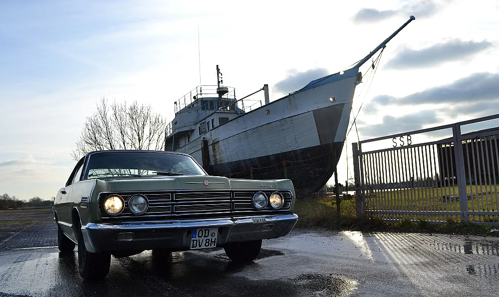 Ein klassisches Auto steht auf einer nassen Straße, mit einem großen, alten Schiff im Hintergrund. Das Auto hat das Nummernschild "OD DV 8H" und die Szene spielt vermutlich am späten Nachmittag oder frühen Abend.