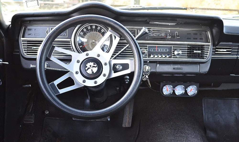 Das Bild zeigt das Armaturenbrett eines klassischen Autos mit einem Dreispeichen-Lenkrad und einem Tachometer bis 120 mph. Es gibt verschiedene Bedienelemente und Anzeigen, darunter eine analoge Uhr und ein Radio.