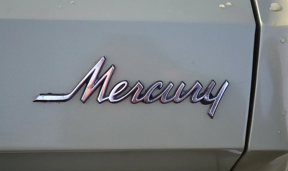 Das Bild zeigt das Emblem eines Mercury-Fahrzeugs. Das Emblem ist in einer kursiven Schrift und metallisch, reflektiert das Licht. Der Hintergrund scheint die Karosserie des Autos zu sein, die in einer hellen Farbe lackiert ist.