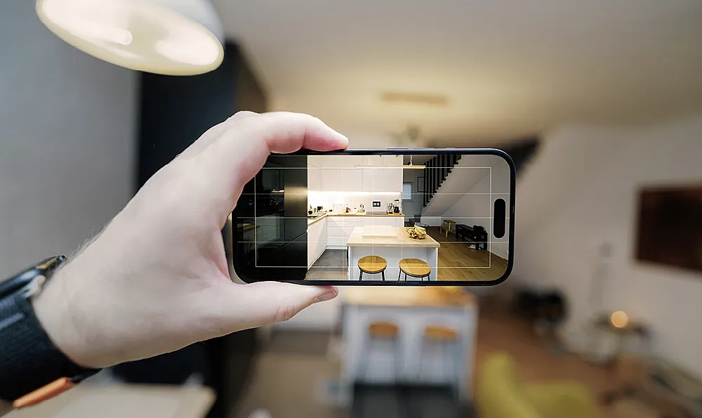 Eine Hand hält ein Smartphone, das eine stilvolle Küche mit eleganten schwarzen und weißen Schränken und hölzernen Barhockern erfasst. Die Szene strahlt Modernität und schickes Design aus.