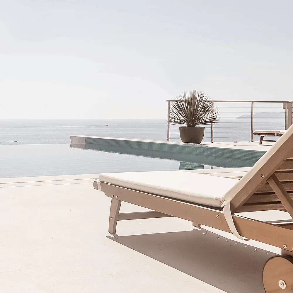 Sonnenliege aus Holz steht an einem Infinity-Pool mit Pflanze und Meer im Hintergrund.