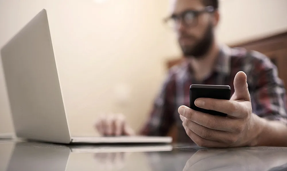 Eine Person sitzt an einem Schreibtisch, benutzt einen Laptop und hält ein Smartphone. Das Gesicht der Person ist unscharf, und der Fokus liegt auf den verwendeten Geräten.