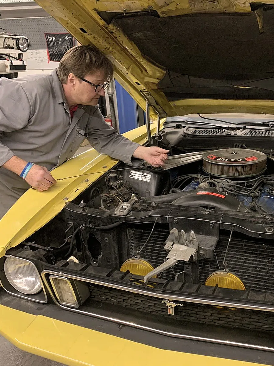 Ein Mechaniker in grauer Arbeitskleidung arbeitet an einem gelben Oldtimer mit geöffneter Motorhaube.