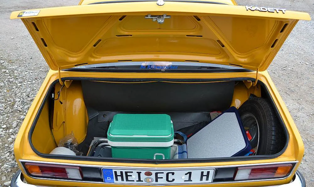Offener Kofferraum eines gelben Opel Kadett mit einer grünen Kühlbox, einer grauen Tasche und einem Ersatzreifen.