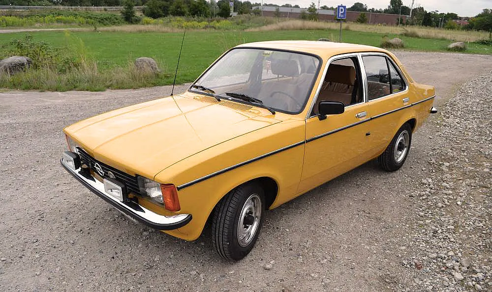 Ein gelbes Vintage-Auto, vermutlich ein Opel Kadett aus den 1970er Jahren, steht auf einem Kiesweg mit einer Wiese im Hintergrund.