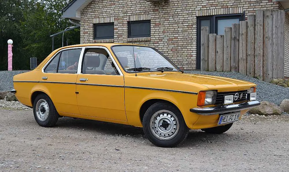 Gelbes Vintage-Auto auf einem Kiesplatz vor einem Backsteingebäude mit Holzzaun.