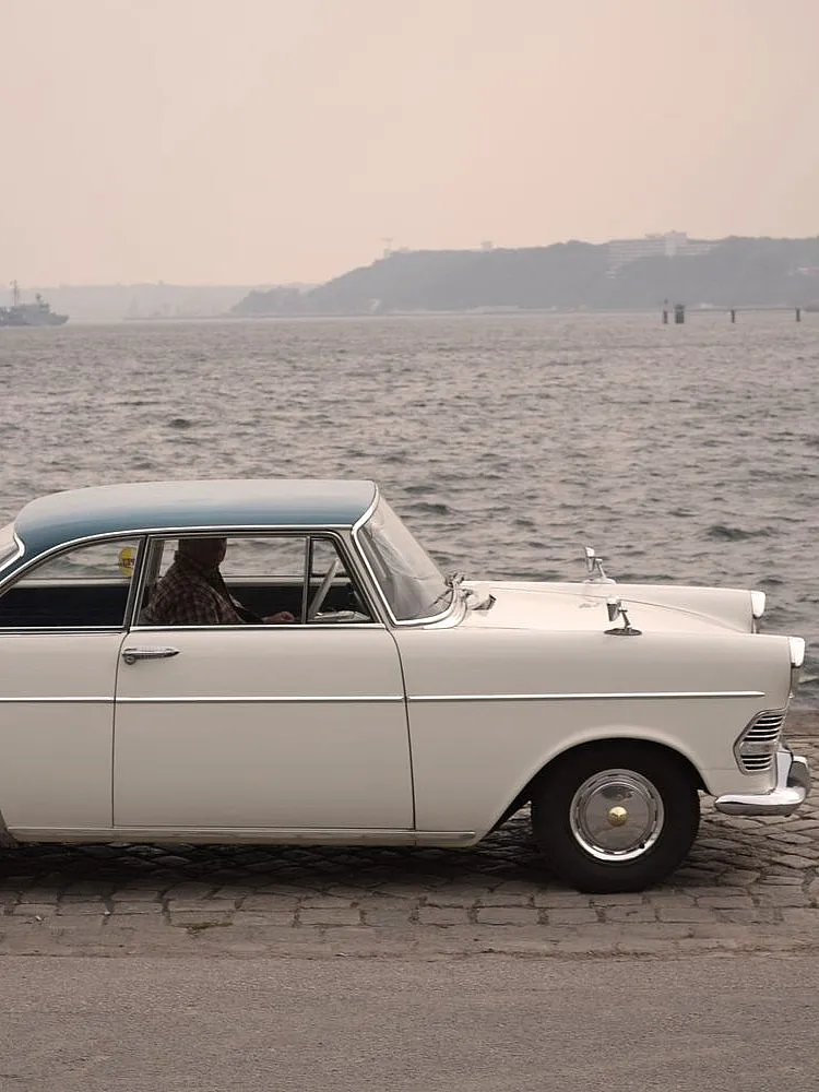 Weißes klassisches Auto mit blauem Dach und Chromdetails, geparkt auf Kopfsteinpflaster nahe einem Gewässer.