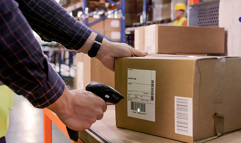 Paket liegt auf einem Tisch in einem Warenlager. Ein Mann in dunkelblauem Hemd hält einen Scanner in der rechten Hand und scannt das Paket en.