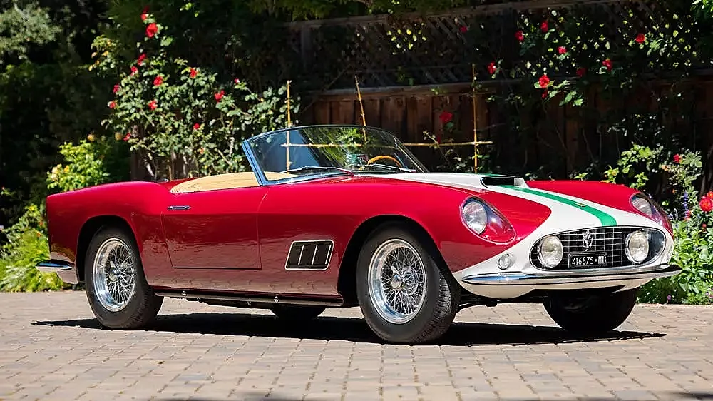 Rotes Ferrari mit weißen und grünen Streifen auf der Motorhaube, geparkt auf Pflastersteinen vor einer Hecke mit blühenden Rosen.