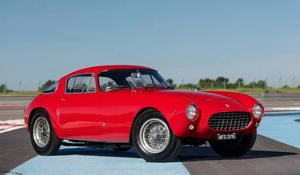 Klassischer roter Sportwagen mit aerodynamischem Design und Speichenrädern, geparkt auf einer Rennstrecke.