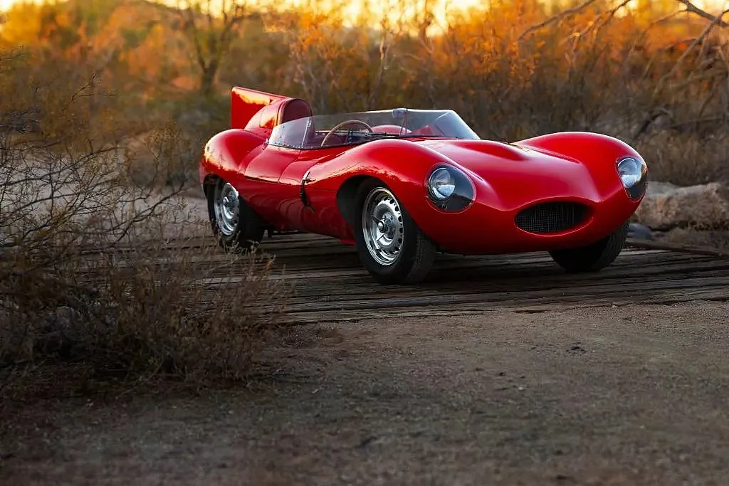Rotes klassisches Sportauto mit offenem Cockpit auf sandigem Weg in Wüstenlandschaft mit trockenen Büschen im Hintergrund.