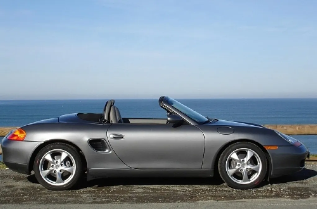 Fotografie eines grauen Cabrio-Sportwagens, seitlich aufgenommen vor Küstenlandschaft mit Meer und blauem Himmel. Fokus auf aerodynamisches Design, offene Dachkonstruktion und glänzende Felgen.