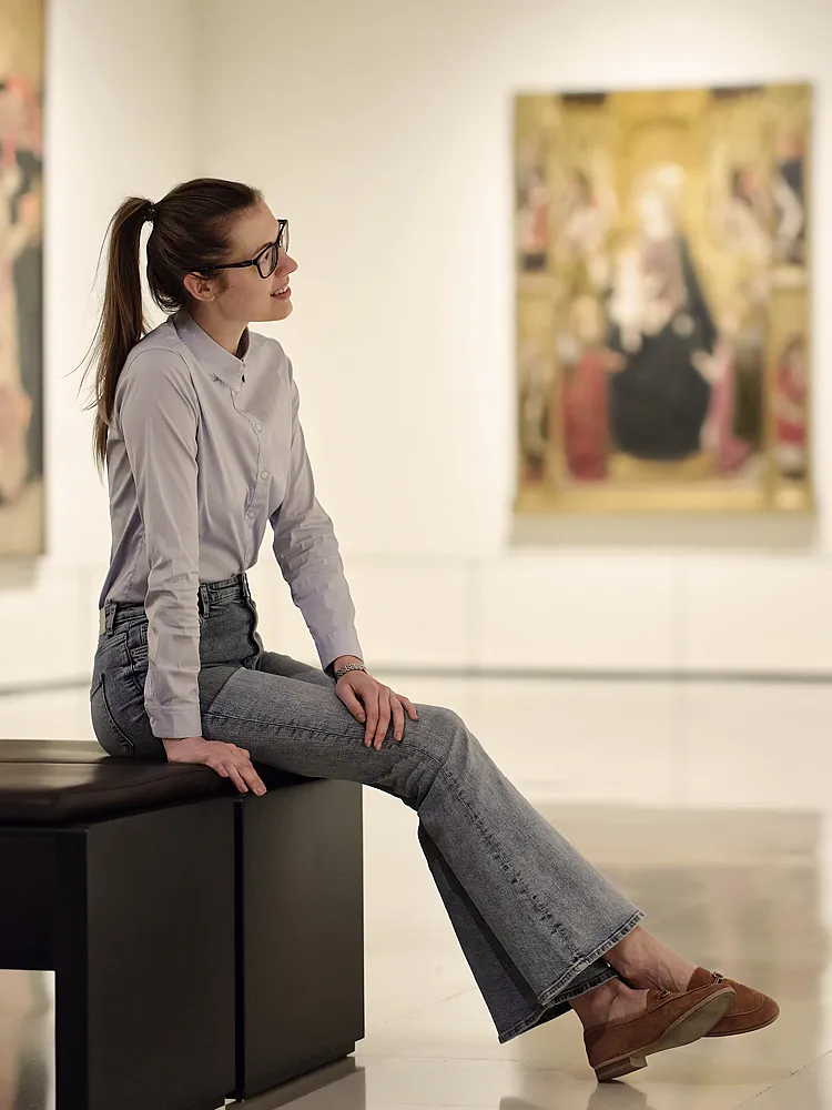 Eine junge Frau sitzt in der Mitte einer Kunstausstellung, an den Wänden hängen Bilder