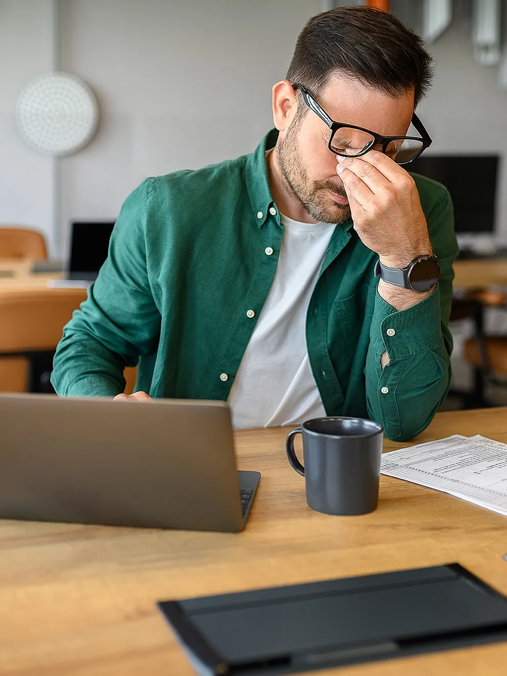 Ein Mann in einem grünen Hemd sitzt an einem Schreibtisch, sieht gestresst aus und drückt sich die Nasenwurzel. Er ist umgeben von Papieren, einem Laptop, einem Tablet und einer Kaffeetasse.