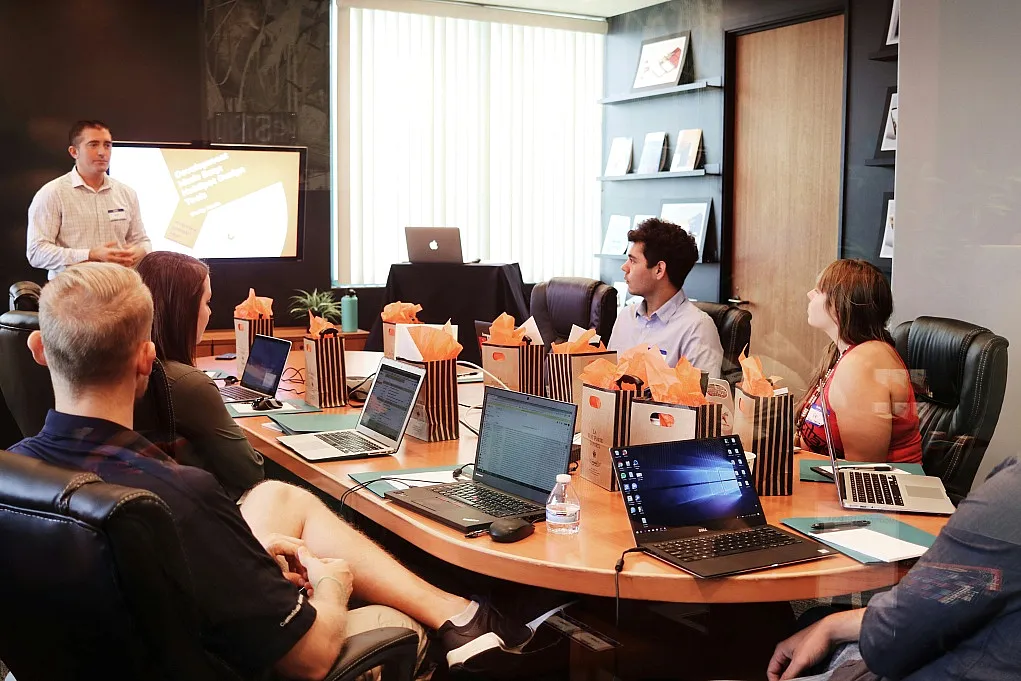 Fotografie einer Geschäftspräsentation in einem modernen Konferenzraum mit sieben Personen, die an einem ovalen Tisch sitzen, auf Laptops arbeiten und einem Mann zuhören, der vor einem Bildschirm steht. Der Bildschirm zeigt eine Folie mit Text und einem Diagramm, während auf dem Tisch mehrere Geschenktüten und Wasserflaschen verteilt sind.
