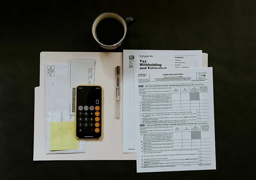 Fotografie zeigt Steuerunterlagen, einen Taschenrechner auf einem Smartphone, einen Stift und eine Tasse Kaffee auf dunklem Untergrund. Fokus liegt auf Formularen zur Steuerabzugs- und Erstattungsanmeldung, was auf Vorbereitung oder Bearbeitung von Steuererklärungen hinweist.
