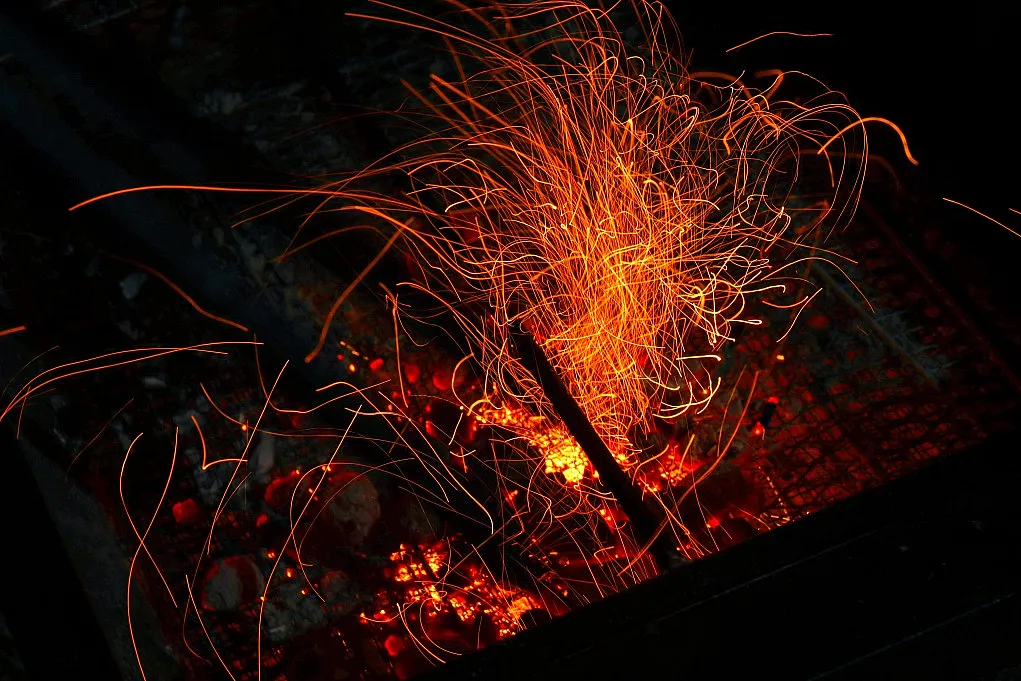 Fotografie eines glühenden Lagerfeuers mit Funkenflug, die dynamische Bewegung und Hitze visualisiert. Dunkler Hintergrund hebt orange-rote Funken und glühende Kohlen hervor, was lebendige Energie und Wärme vermittelt.