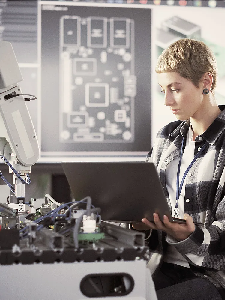 Eine junge blonde Frau mit kurzen Haaren sitzt vor einer Werkbank mit elektronischen Geräten und einem Roboterarm. Sie hält einen Laptop in der Hand über ihrem Schoß, in den sie hineinschaut. Im Hintergrund sieht man verschwommen einen großen Bildschirm mit technischen Zeichnungen.