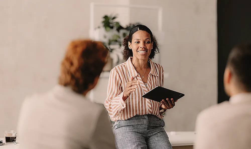 Eine Frau mit einem Tablet unterhält sich mit zwei sitzenden Personen. Sie lächelt, trägt ein gestreiftes Hemd und Jeans, im Hintergrund ist eine Pflanze zu sehen, was eine freundliche und professionelle Atmosphäre vermittelt.