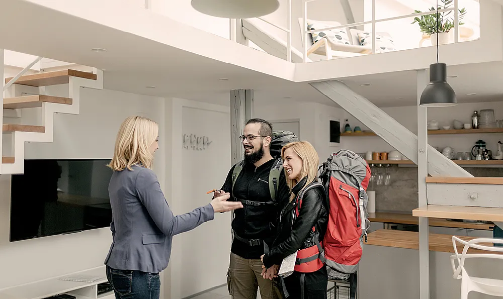 Eine Frau mit blondem Haar übergibt einem lächelnden Paar mit großen Rucksäcken einen Schlüssel für eine moderne Ferienwohnung mit einer Treppe und einem offenen Obergeschoss.